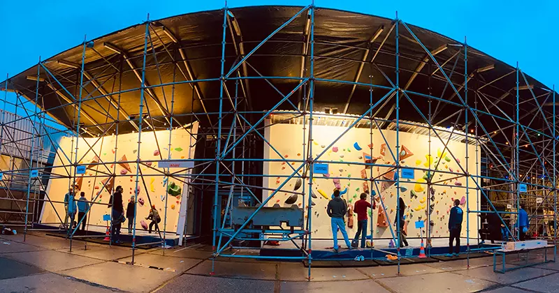 Tijdens een avondevenement in de open lucht zijn mensen aan het boulderen op kunstmatige klimwanden die omringd zijn door steigers. Het tafereel wordt verlicht door kunstlicht en er zijn verschillende klimmers en toeschouwers aanwezig.